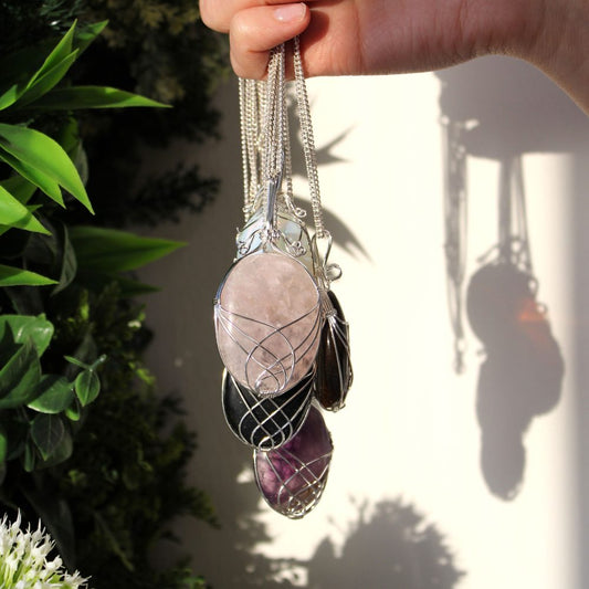 A hand holds several crystal pendants with silver wiring and chains, dangling in front of a white wall and leafy green plants.