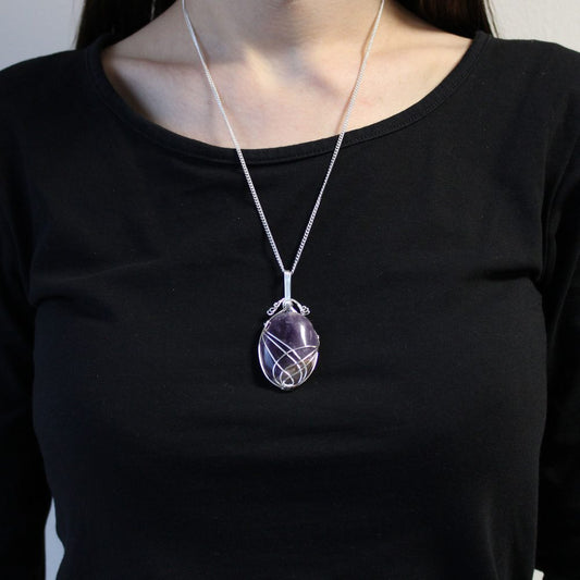 A person wears a silver necklace with a large, oval, purple stone pendant wrapped in silver wire. The person is wearing a black long-sleeved shirt, and the background is a neutral off-white.