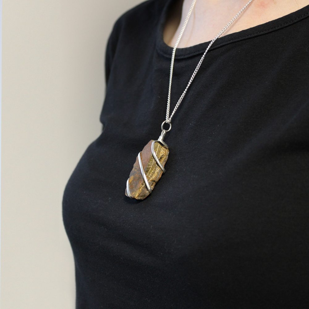 A brown, oblong stone pendant wrapped in silver wire hangs from a silver chain around a person's neck. The person wears a dark-colored shirt against a light background.