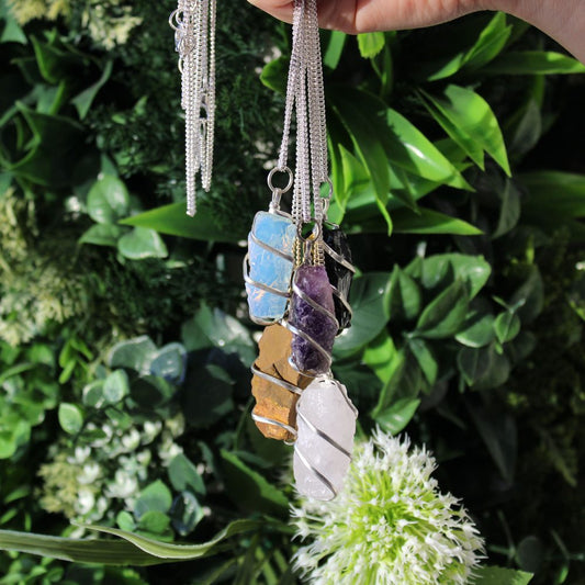 A hand holds five silver chain necklaces with rough, wrapped crystals—opalite, amethyst, tiger's eye, quartz, and black tourmaline—against a backdrop of green foliage and white flowers.