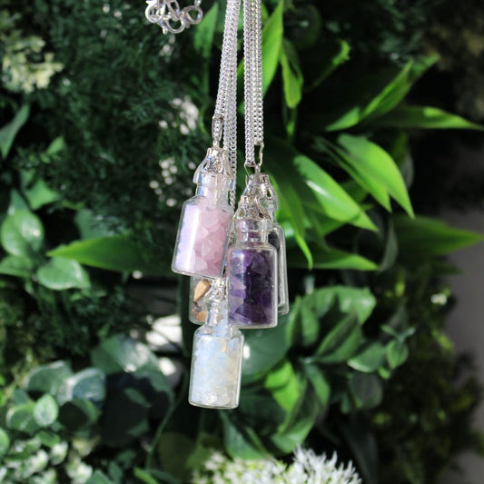 Necklaces with crystal-filled miniature bottles dangle against a lush, green backdrop of leaves and foliage. The crystals within are white, amethyst, rose quartz, and brown colored.