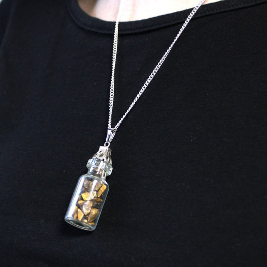 A small glass vial filled with brown stones hangs on a silver chain necklace against a dark-colored shirt. The bottle has an ornate, clear glass cap.
