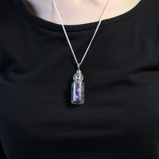 A small glass vial, filled with small purple stones, hangs on a silver chain around a person's neck, set against a plain black shirt.