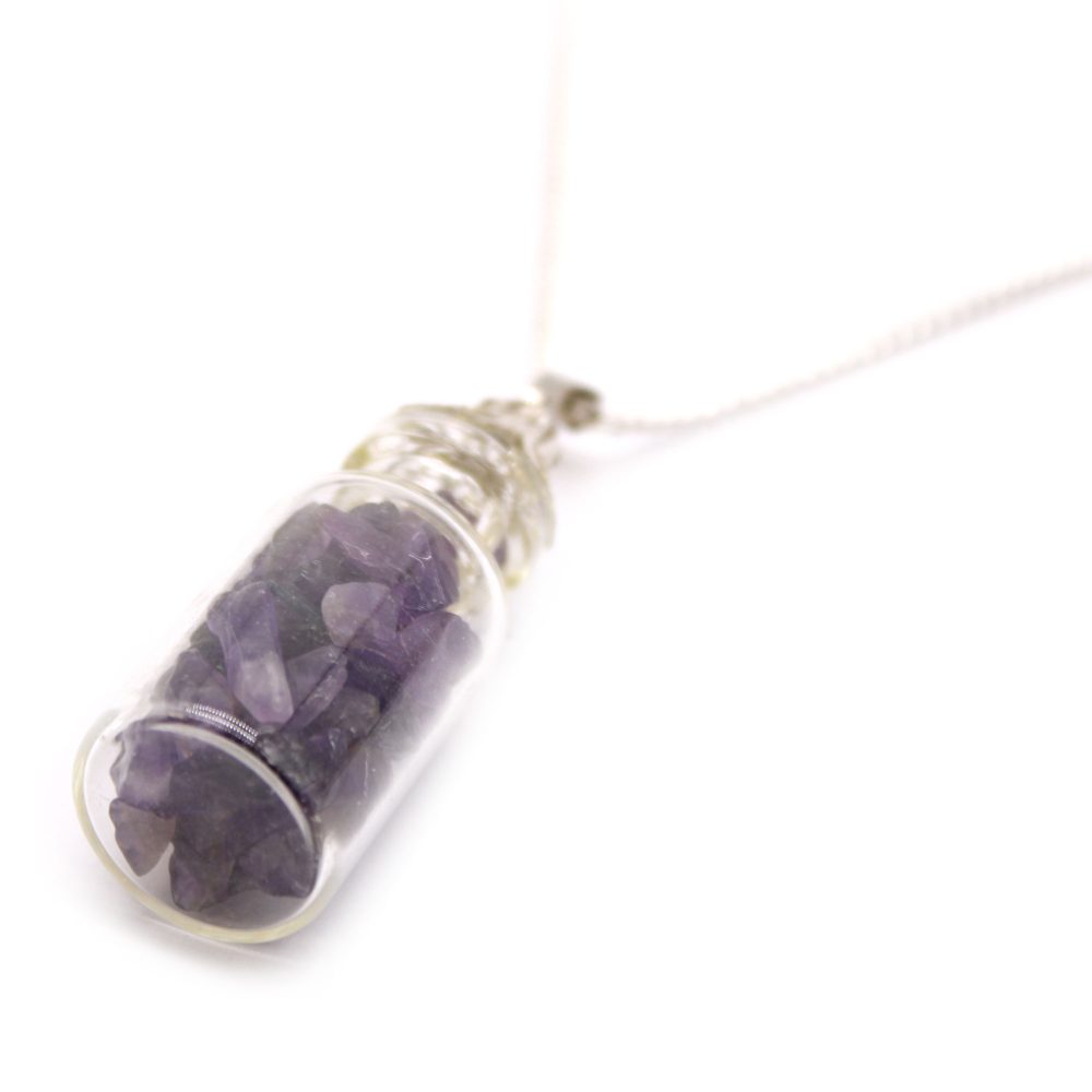 A small glass bottle necklace, filled with purple amethyst crystals, hangs diagonally on a white surface. Its silver chain extends upwards to the right.