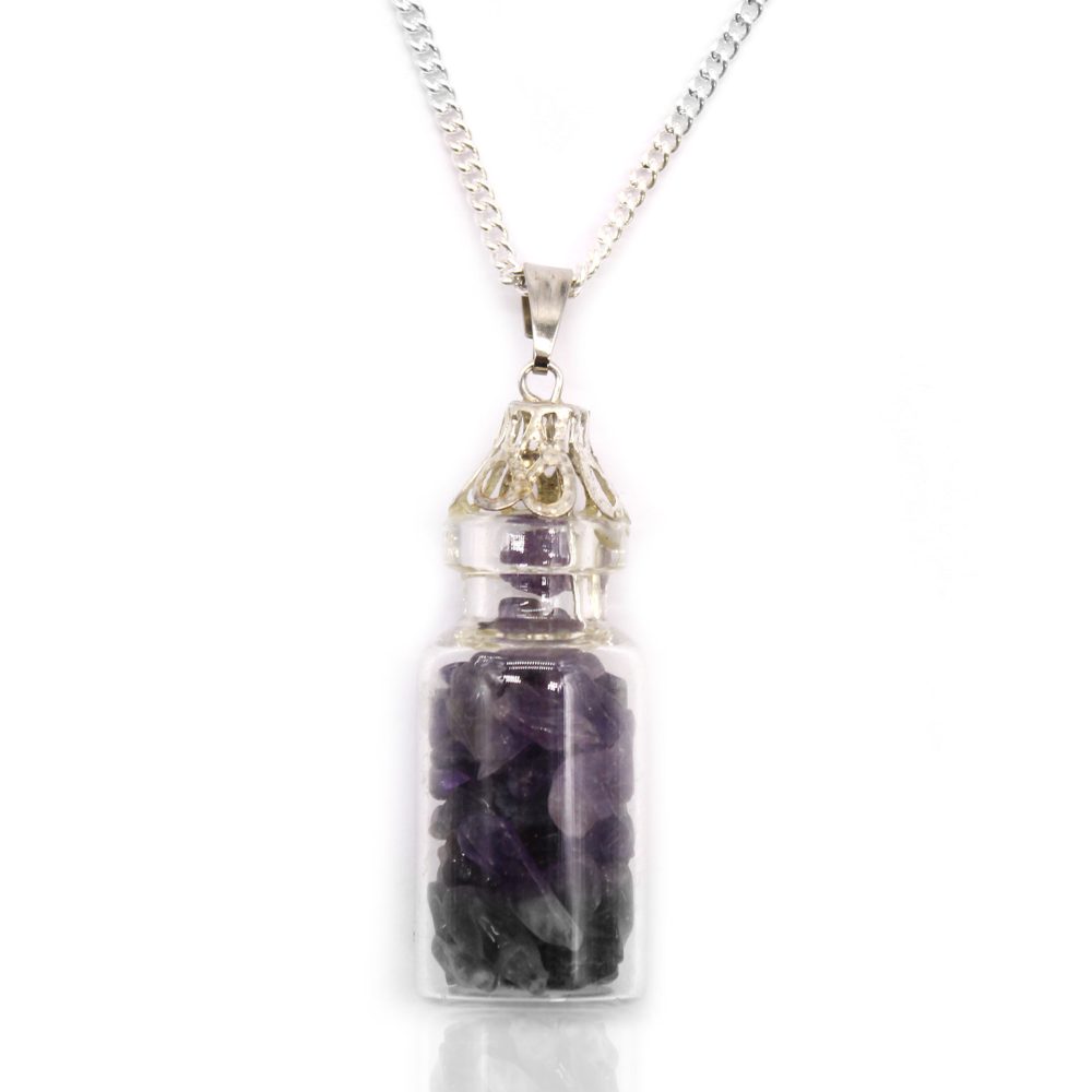A small glass bottle necklace filled with purple and grey stones is suspended from a silver chain. It reflects on a bright white surface.