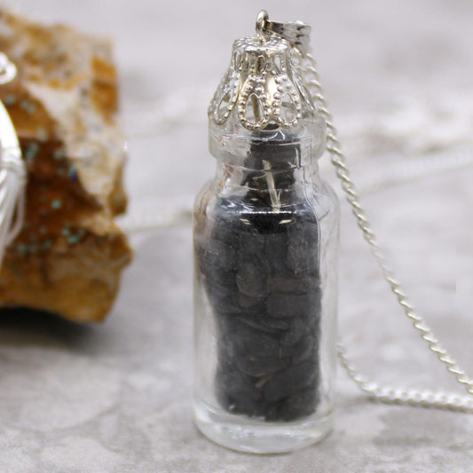 A clear bottle pendant, filled with dark pebbles, hangs from a silver chain. It sits against a mottled gray surface near a rough rock.
