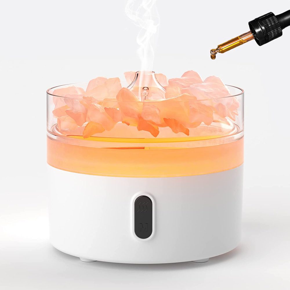 A white, round aroma diffuser emits vapor. The diffuser contains orange liquid, peach-colored crystals, and a dropper is positioned to drip golden oil in. The diffuser sits on a white surface.