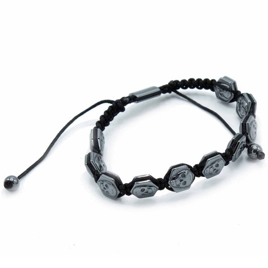 A black woven bracelet features metal hexagon charms with skull carvings. The bracelet lies on a plain white surface.