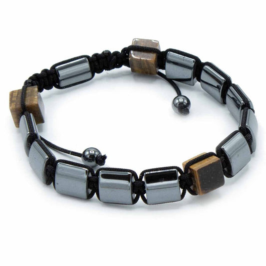 A bracelet is displayed on a white surface. It’s made of black cord threaded through alternating square beads of grey hematite and tan tiger's eye.