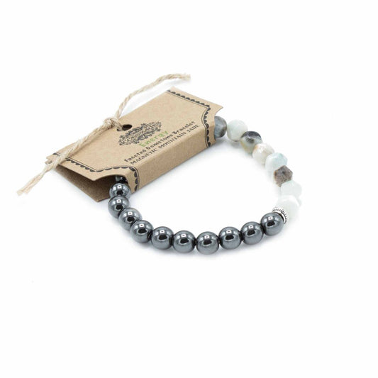 A gemstone bracelet, with an attached tag bearing the text "Faceted Gemstone Bracelet Energy BY MAGNETIC MOUNTAIN JADE", rests on a white surface. The bracelet alternates between dark, metal-colored beads and lighter, semi-transparent stones.