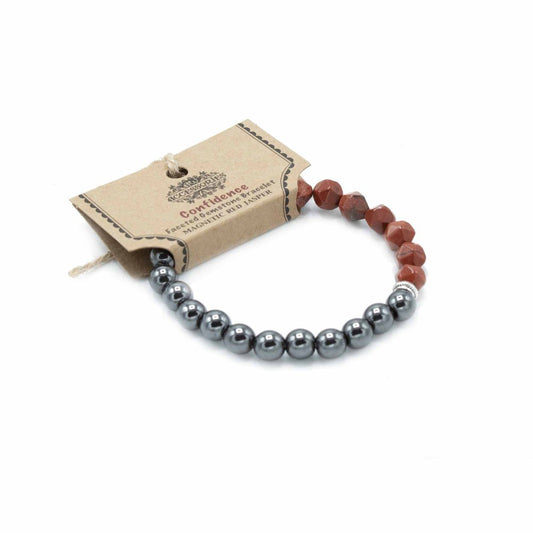 A bracelet, composed of metallic gray spheres and red jasper beads, rests next to a brown tag, reading "Confidence Faceted Gemstone Bracelet MAGNETIC RED JASPER", on a white surface.