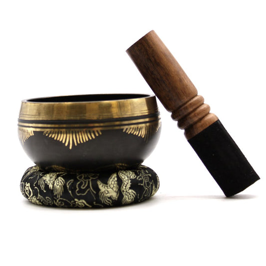 A bronze and black singing bowl rests on an embroidered cushion, accompanied by a wooden striker leaning against it, placed on a plain white background.