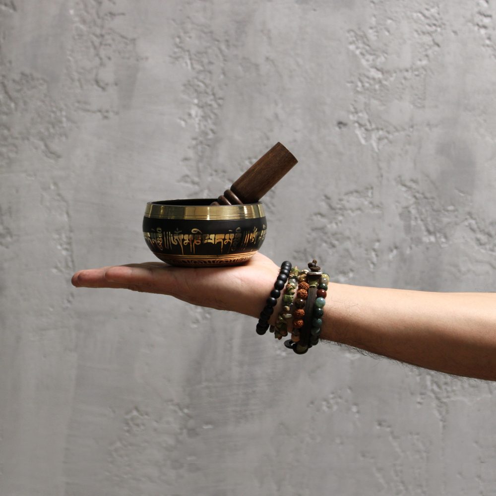 A Tibetan singing bowl with inscribed text rests on an outstretched hand adorned with colorful beaded bracelets, set against a textured gray wall.