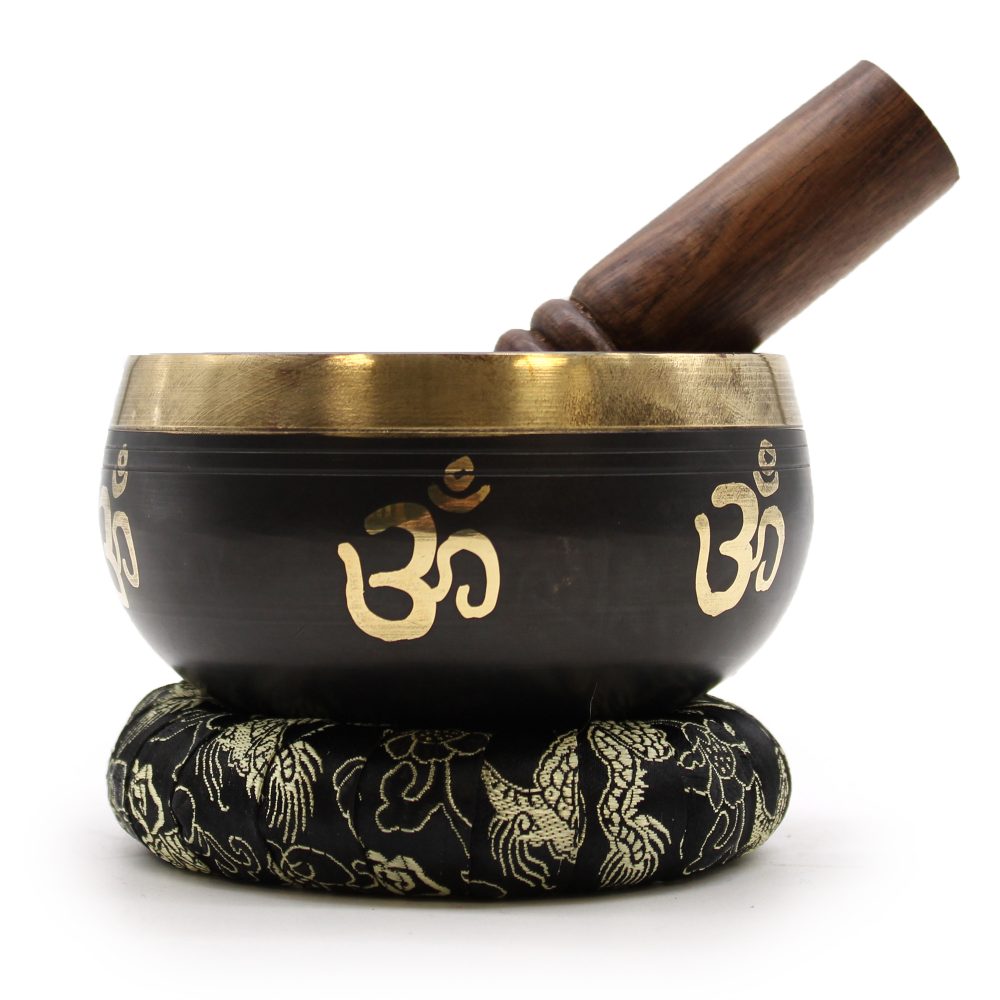 A black and gold singing bowl with "Om" symbols rests on an ornate cushion, with a wooden striker leaning against it.