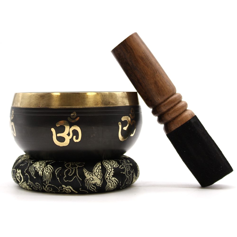 A singing bowl with gold "Om" symbols rests on an intricately patterned cushion. Beside it, a wooden mallet leans against the bowl on a plain white background.