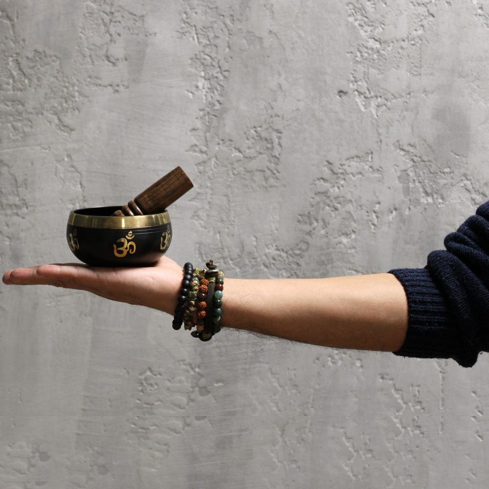 A hand, adorned with beaded bracelets, holds a decorative singing bowl with a wooden mallet. The bowl displays golden "Om" symbols. The setting is a textured gray wall.