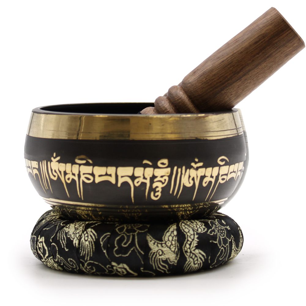 A black and gold singing bowl, etched with intricate script, rests on an ornate cushion. A wooden striker leans inside, highlighting the elegant design and tranquil setting.