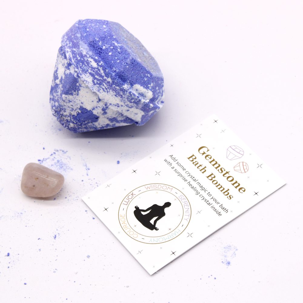 Blue bath bomb resting on white surface, with scattered powder nearby; accompanied by a polished stone and card reading: "Gemstone Bath Bombs. Add some crystal magic to your bath with a surprise healing crystal inside. COURAGE LUCK WISDOM SERENITY HARMONY."