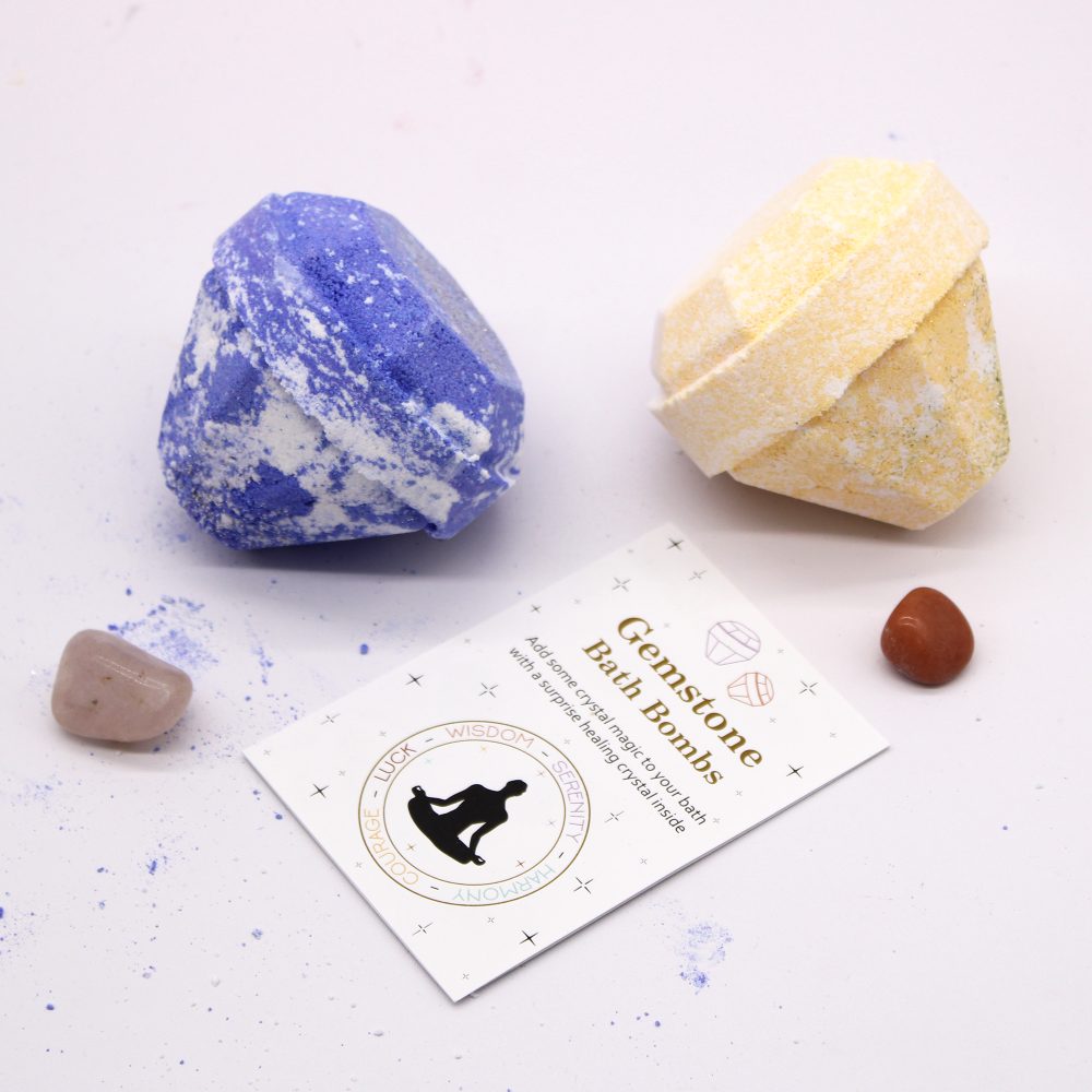 Two gemstone-shaped bath bombs, one blue and one yellow, lie beside small crystals and a card reading: "Gemstone Bath Bombs - A hidden crystal for your bath with surprise healing crystals inside."