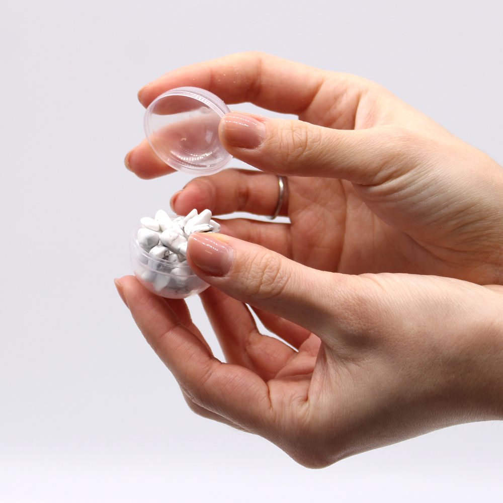 Hands hold an open clear container filled with white tablets, removing the lid. The background is plain and light, focusing attention on the hands and container.