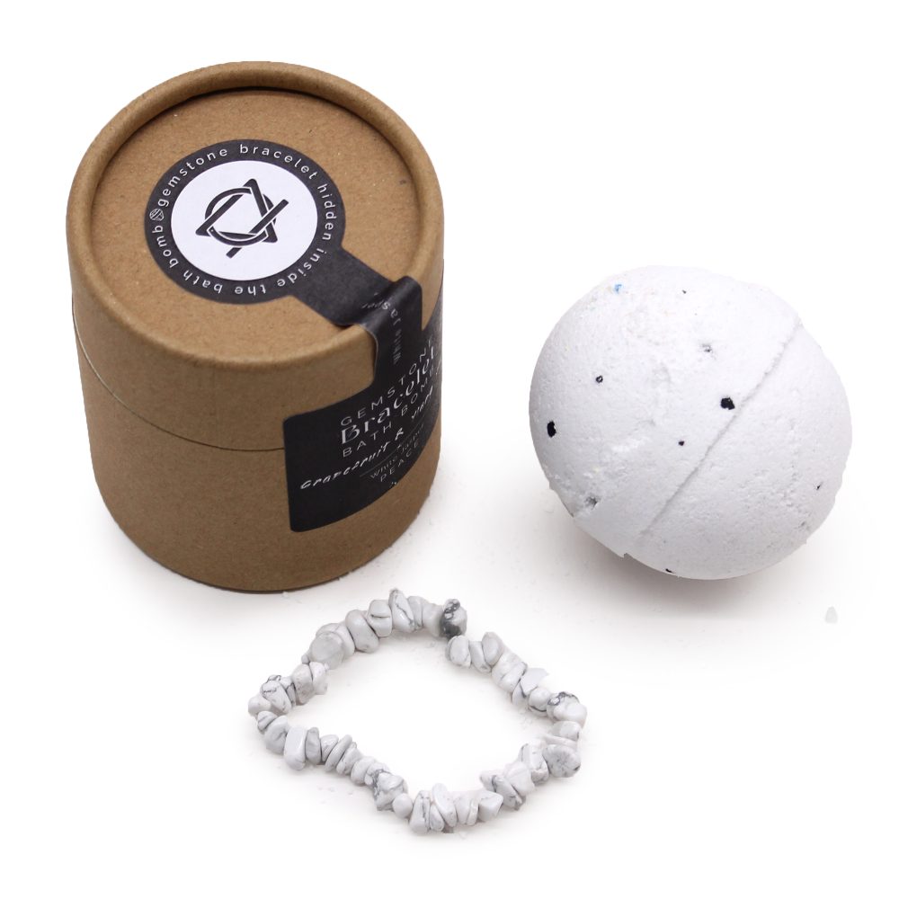 A white bath bomb with black speckles and a gemstone bracelet lie next to a brown cylindrical box labeled: "gemstone bracelet hidden inside the bath bomb," "GEMSTONE DISCOVERY BATH BOMB," "Bracelet with a Purpose!"