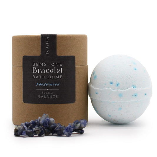A round bath bomb with blue speckles sits beside a brown cylindrical package labeled "GEMSTONE Bracelet BATH BOMB Sandalwood x Sodalite BALANCE." Nearby are scattered blue sodalite stones.