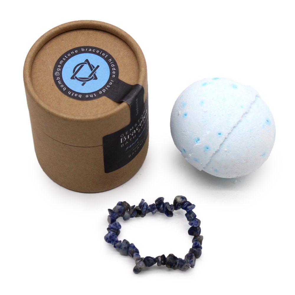 A brown cylindrical box labeled "Gemstone Bracelet Hidden Inside the Bath Bomb," a blue-speckled bath bomb, and a dark beaded bracelet lie on a white surface.