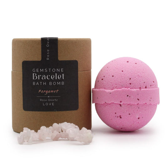 A pink bath bomb sits beside a brown cylindrical package labeled "GEMSTONE Bracelet BATH BOMB" with text "Bergamot, Rose Quartz, LOVE." A gemstone bracelet rests in front.