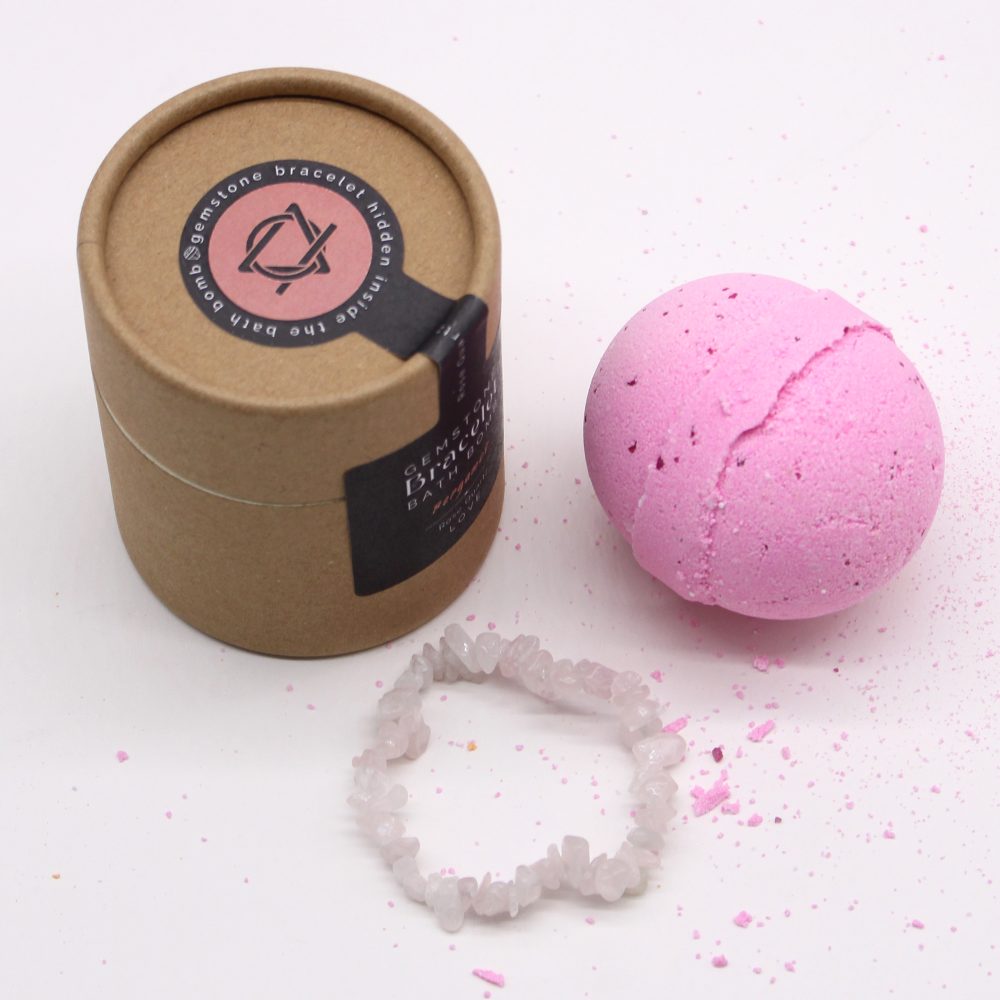 A pink bath bomb with a gemstone bracelet is next to a brown cylindrical container labeled "Hidden gemstone bracelet inside the bath bomb" on a white surface.