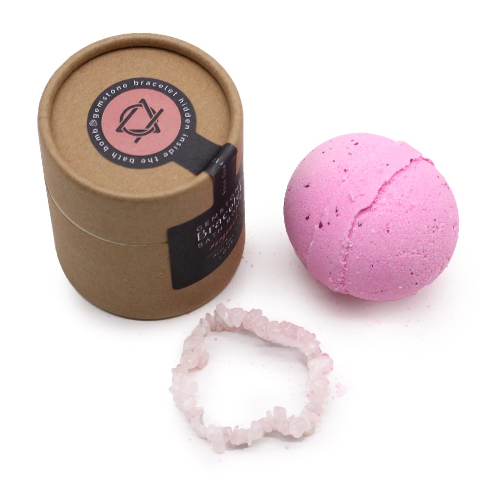 Pink bath bomb sits beside a cardboard container labeled "gemstone bracelet hidden inside the bath bomb," with a gemstone bracelet resting in front. All items are on a white surface.