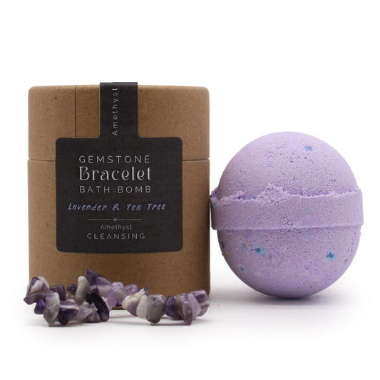 A purple bath bomb rests beside a cylindrical cardboard box labeled, "Gemstone Bracelet Bath Bomb, Lavender & Tea Tree, Amethyst, Cleansing." An amethyst bracelet lies near the box.