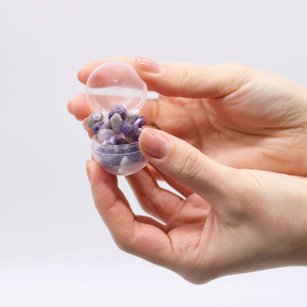 A hand holds an open, transparent capsule containing small purple and white stones, set against a plain white background.