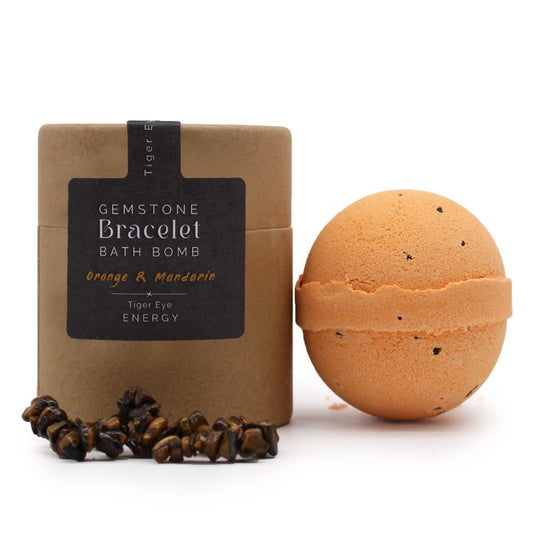 A round orange bath bomb with black specks sits beside a cylindrical box labeled, "Gemstone Bracelet Bath Bomb, Orange & Mandarin, Tiger Eye Energy." A tiger eye gemstone bracelet lies in front.