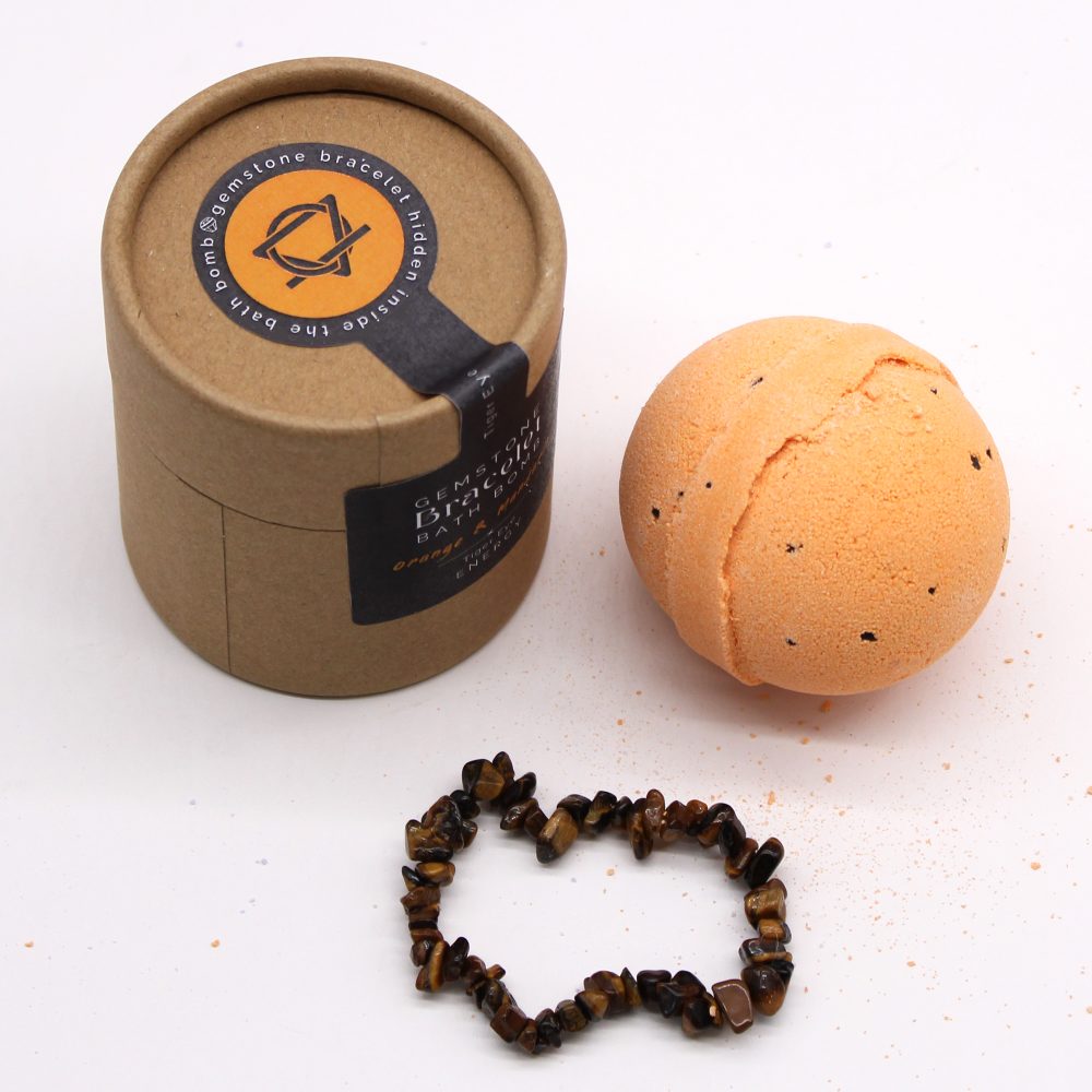 A round, orange bath bomb with black speckles sits beside a brown container labeled "Gemstone Bath Bomb," alongside a gemstone bracelet on a white surface.