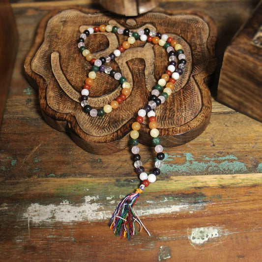 A mala necklace, with colorful beads and tassel, rests atop a carved wooden "Om" symbol placed on a weathered wooden surface.