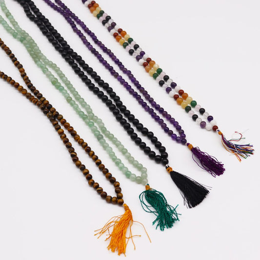 Six mala necklaces lay diagonally across a white surface, each with a different colored strand of beads and matching tassel.