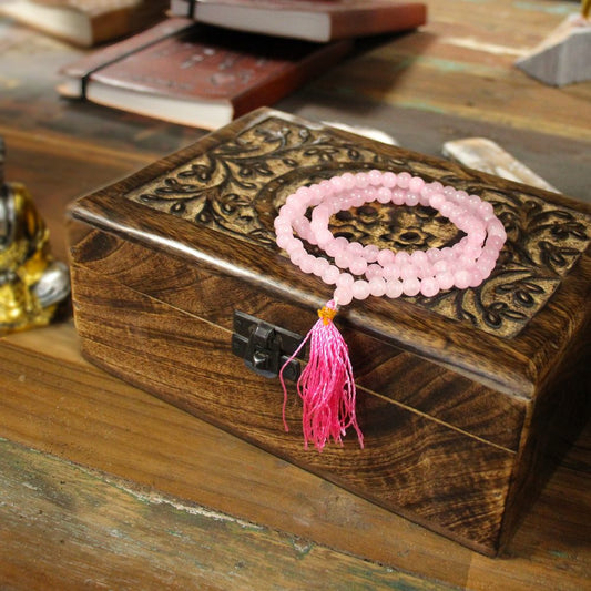 A rose quartz mala rests atop an ornate wooden box with a metal clasp on a weathered wooden table.