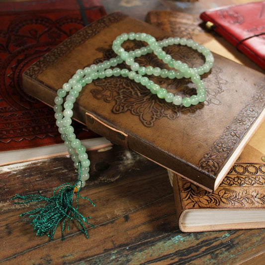Green beaded prayer beads rest on top of a tooled leather journal. The scene is surrounded by other leather journals on a rustic wooden surface.