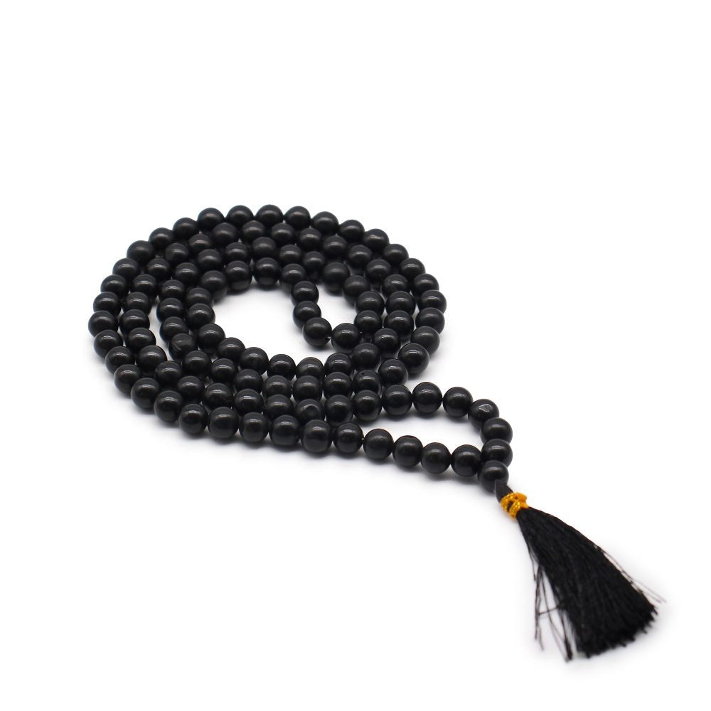 A black beaded mala with a tassel rests on a white surface, the beads are arranged in a coiled shape.
