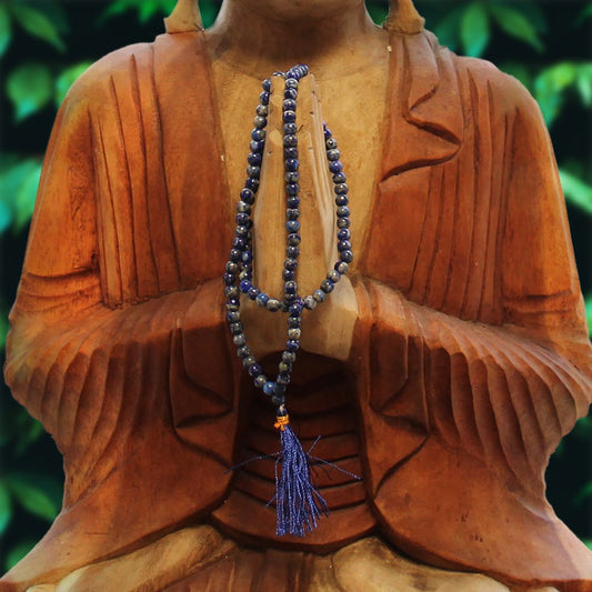 A wooden Buddha statue has its hands clasped together in prayer, with a string of blue beads draped over its hands, against a background of green foliage.
