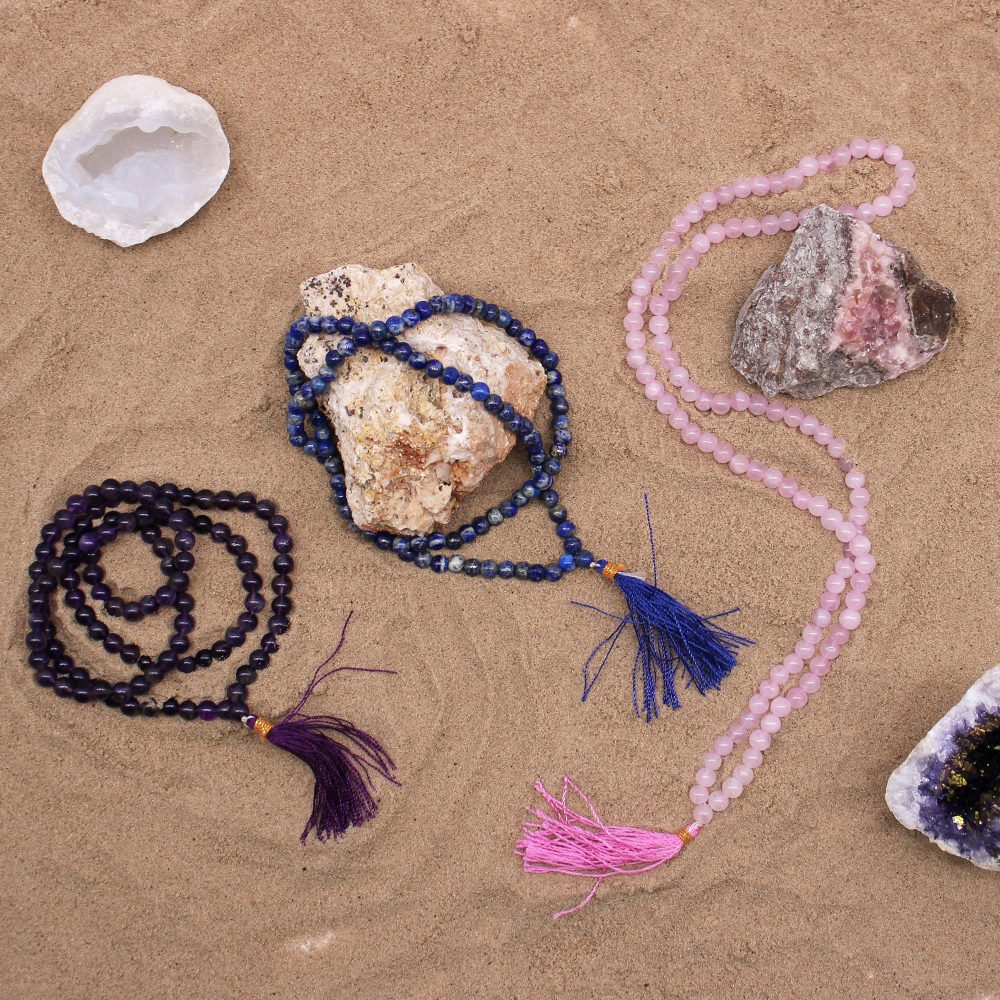 Three beaded necklaces are laid on sand near rocks and geodes. They are purple, blue, and pink, each with a tasseled end.