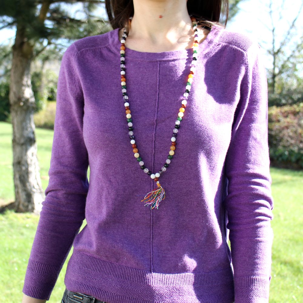 A woman is wearing a long, beaded necklace that has a colorful tassel, outdoors in a grassy area with a tree in the background.
