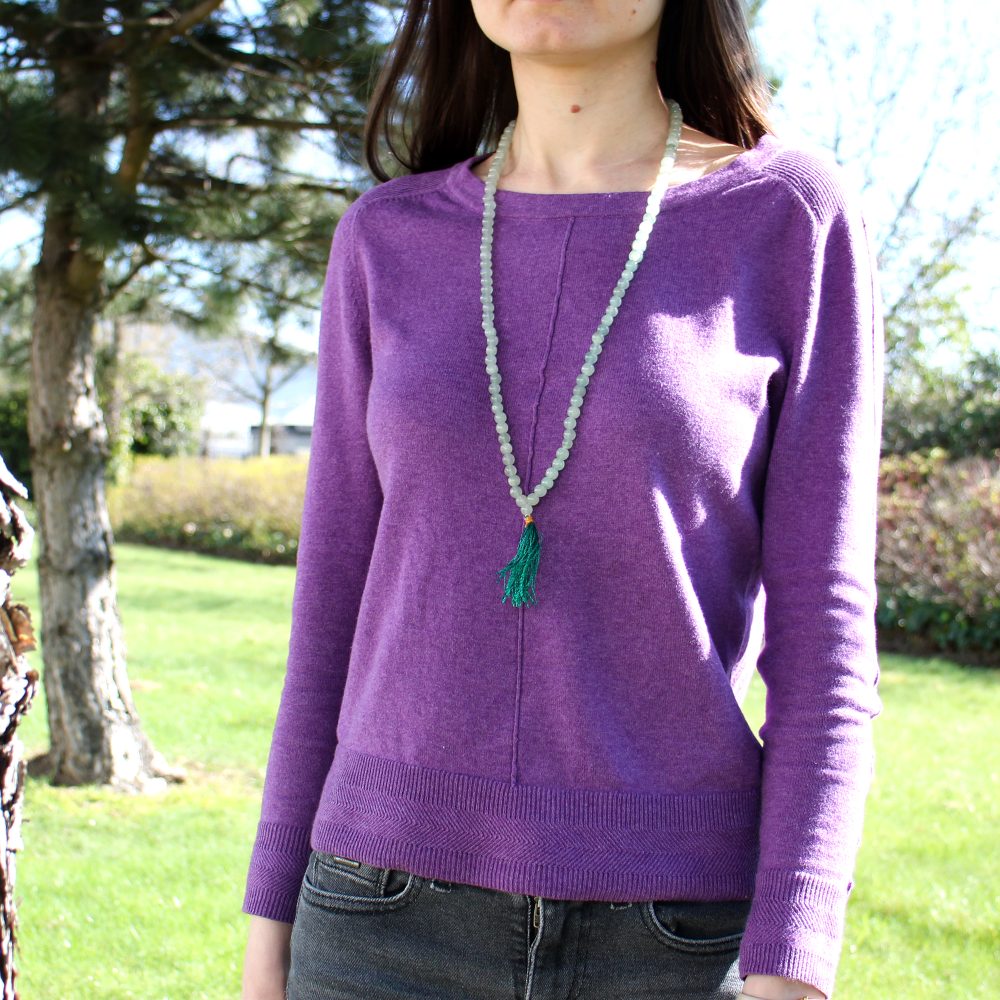 A woman stands in a park setting. She wears a purple sweater and a green beaded necklace with a tassel.