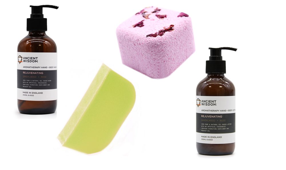 Two amber bottles of Ancient Wisdom Rejuvenating Sandalwood & Rose hand wash and lotion, a light green soap, and a pink bath bomb sit on a white background.