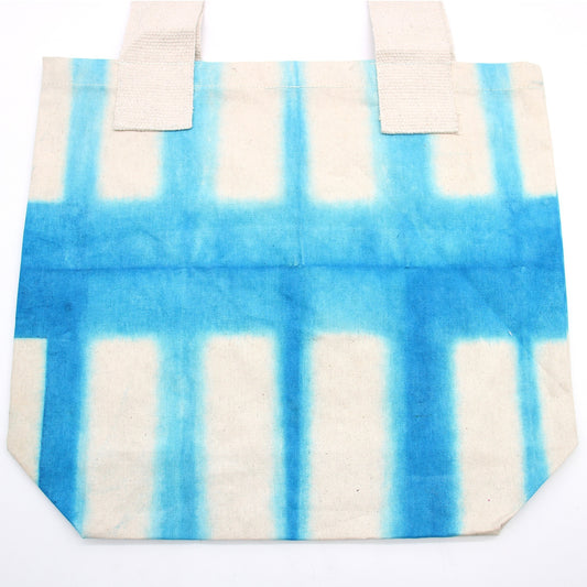 A white canvas tote bag with two handles displays a tie-dye pattern of light blue stripes forming a grid on a white background. The bag is on a white surface.