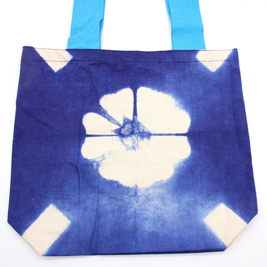 A tote bag sits upright. It features a large, white floral design on a blue background with light blue handles and is on a white surface.