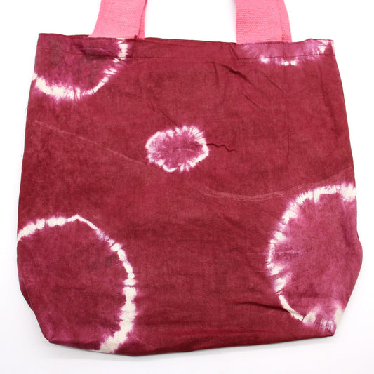 A burgundy tote bag with pink handles sits on a white surface. The bag features white tie-dye circle patterns.