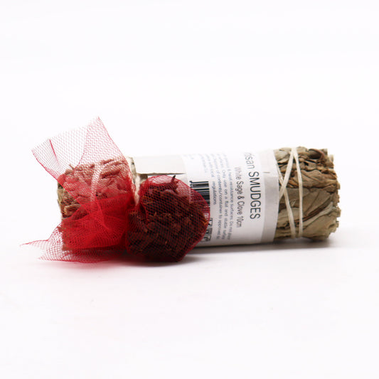 A sage and clove smudge stick rests on a white surface; it's bundled with twine and adorned with a red mesh bag of cloves.