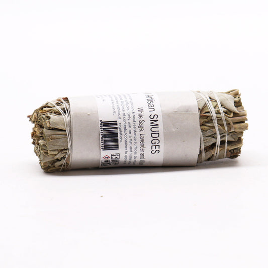 A smudge stick of white sage, lavender, and mugwort rests on a white surface. The stick is bound with twine and wrapped in paper with product information.