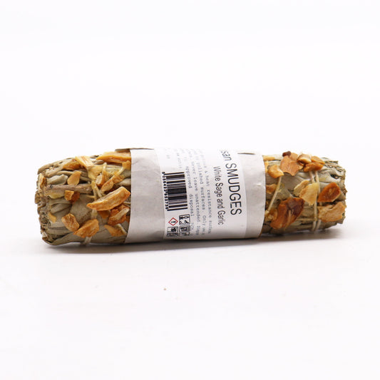 A sage smudge stick, containing white sage and garlic pieces, rests on a white surface.